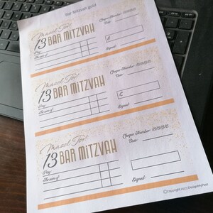 May include: A white sheet of paper featuring three printed templates for Bar Mitzvah gift cheques. Each template includes the text Mazel Tov! 13 Bar Mitzvah, with fields for cheque number, date, amount, payee, and signature, accented with gold glitter.