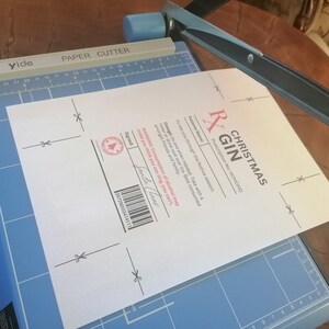 May include: A white card with a black and pink "Christmas Gin" prescription label. The label includes the text "Prescription No: HOHOHO" and "To see you through the festive season." The card is on a blue cutting mat with a paper cutter.