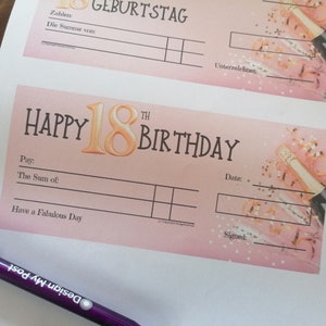 Two pink and gold birthday gift cheques with the text 'Happy 18th Birthday' and a gold number 18 balloon. The check has a space to write the amount of money given and a space to sign the card. This design is in German and in English. The cheque has a pink background with gold confetti.