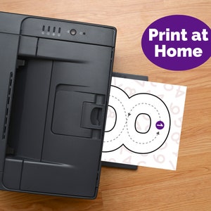 May include: A black desktop printer outputting a white sheet of paper featuring a large black outline of the number eight with directional arrows and a purple circle containing the number one. A purple oval with the text Print at Home is visible.