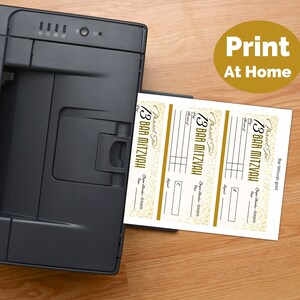 May include: A black printer feeding a sheet of paper with three printed Bar Mitzvah check templates. Each template features gold glitter accents, the text "Mazel Tov!", "BAR MITZVAH", "Pay", "Date", "Check Number", and "Signature". A gold circle with "Print At Home" is visible.
