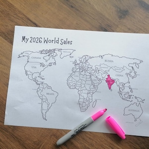 May include: A white paper sheet featuring a black outline world map with the text My 2026 World Sales at the top. The country of India is colored in bright pink. A gray permanent marker with a pink cap is positioned diagonally across the bottom.