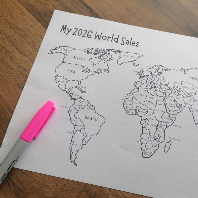 May include: A white paper sheet featuring a black and white printed world map with the title My 2026 World Sales at the top. A bright pink felt-tip marker with a gray body is positioned diagonally across the lower left corner of the map.