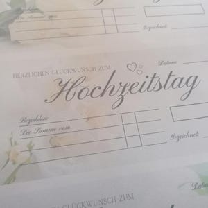May include: Close-up of a light-colored paper with the German phrase "HERZLICHEN GL&Uuml;CKWUNSCH ZUM Hochzeitstag" in elegant script. The paper has lines for filling in details and the word "Datum" is visible. Soft floral accents are in the background.