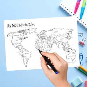 May include: A white sheet of paper featuring a black and white outline map of the world with the text My 2026 World Sales printed at the top. The map includes labeled continents and countries, with a black pen positioned over the African continent.
