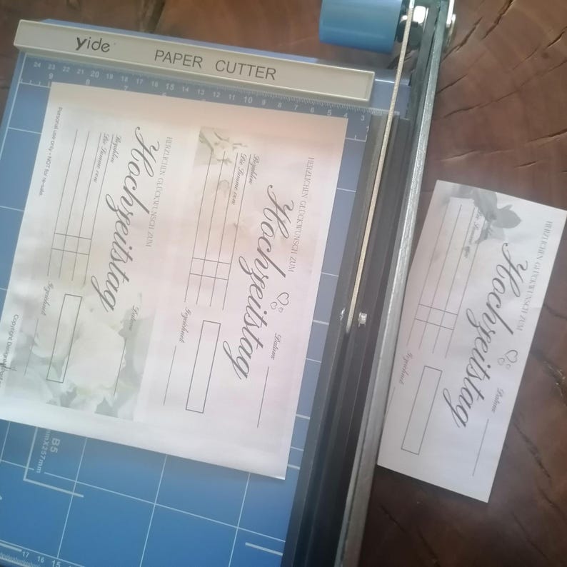 May include: A blue paper cutter with a white label that reads "PAPER CUTTER" is cutting a sheet of paper. The paper has the word "Hochzeitstag" written on it in cursive. A cut piece of paper is to the right.