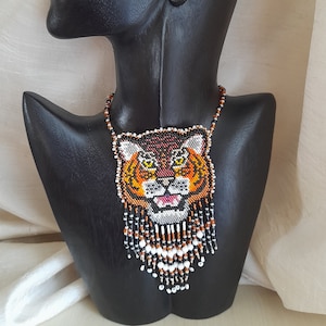 Puede incluir: Un collar de cuentas con un colgante de cara de tigre. El colgante está hecho de cuentas de color naranja, negro, blanco y marrón. El collar tiene una franja de cuentas que cuelgan de la parte inferior.