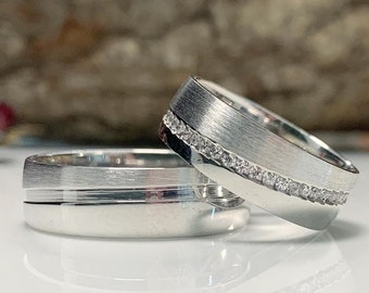 May include: Two silver wedding bands, one with a row of small diamonds. The bands are on a white surface.