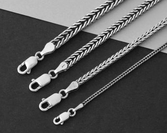 May include: Four silver chains of varying lengths, with a box chain design. The chains are laid out on a gray surface.