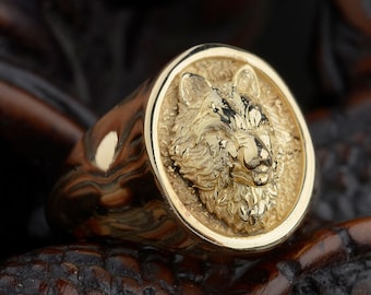 May include: A gold ring with a detailed wolf head design. The ring is set against a dark brown wooden background.