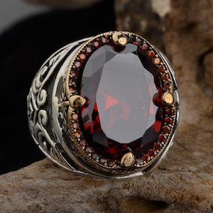 May include: A silver ring with a large, oval-shaped, dark red gemstone surrounded by smaller, clear gemstones. The ring has an intricate, ornate design.