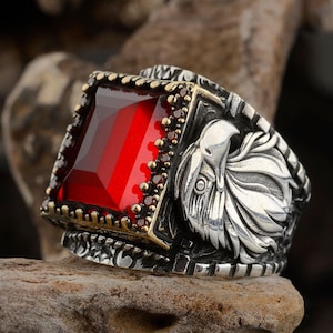 Könnte beinhalten: Ein silberner Ring mit einem großen, rechteckigen roten Edelstein. Der Ring hat ein Adlerdesign an der Seite und einen goldfarbenen Rand um den roten Stein. Der Ring liegt auf einem Holzstück.