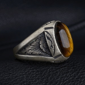 May include: A silver ring with an oval, amber-colored tiger's eye stone. The ring features engraved details on the sides, including a sunburst design. The ring is set against a dark background.