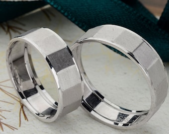May include: Two silver wedding bands with a geometric, faceted design. The rings are displayed on a light-colored surface with a green ribbon and a decorative frame in the background. The rings have a matte finish.