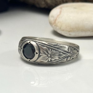 Op de afbeelding: Een zilveren ring met een zwarte edelsteen in het midden. De ring heeft gedetailleerde bloemen- en bladgravures langs de band. De ronde edelsteen is gezet in een bezel. De ring ligt op een wit oppervlak.