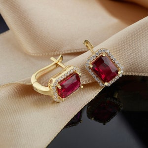 May include: A pair of gold hoop earrings featuring a rectangular-cut deep red gemstone at the center. Each stone is surrounded by a halo of small, clear white pavé-set stones, creating a bright contrast against the polished gold metal setting.