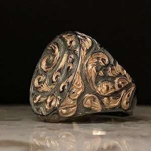 May include: A silver ring with intricate gold filigree detailing. The ring features a rounded face with an ornate, swirling pattern. The design is a mix of silver and gold, creating a striking contrast. The ring is likely a piece of jewelry.