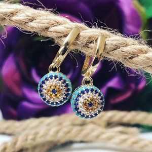 Könnte beinhalten: Ein Paar goldene Creolen mit einem blauen und türkisfarbenen bösen Blick-Anhänger, der an jeder Creole baumelt. Die Anhänger sind mit kleinen weißen, blauen und gelben Steinen besetzt.