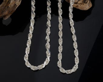 May include: Two silver rope chains, one longer than the other, are displayed on a wooden surface. The chains are made of a twisted rope design.