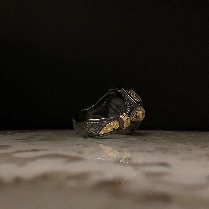 May include: A detailed, dark silver ring with gold accents. The ring features intricate floral and leaf patterns, with a prominent gold teardrop-shaped design. The ring is set against a dark background, highlighting its ornate details.