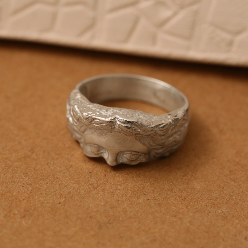 K&ouml;nnte beinhalten: Ein silberner Ring mit einem detaillierten Gesichtsdesign. Das Gesicht ist mit einer detaillierten Frisur und Gesichtsz&uuml;gen gestaltet.