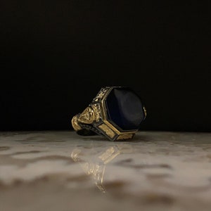 May include: A gold and silver ring with a large, dark blue, octagonal stone. The ring has intricate gold detailing around the stone and on the band. The ring is set against a dark background.