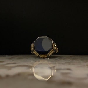May include: A gold-toned ring with an octagonal, dark blue stone. The ring has intricate detailing around the stone and on the band. The ring is displayed on a reflective surface against a dark background.