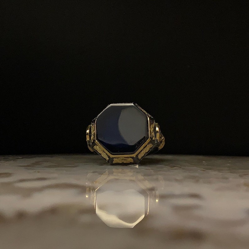 May include: A gold-toned ring with an octagonal, dark blue stone. The ring has intricate detailing around the stone and on the band. The ring is displayed on a reflective surface against a dark background.