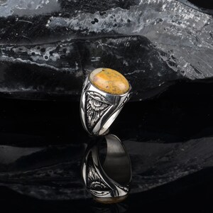Peut inclure: Une bague en argent avec une pierre ovale jaune et brune sertie dans un entourage en argent d&eacute;taill&eacute;. La bague est sur un fond noir.