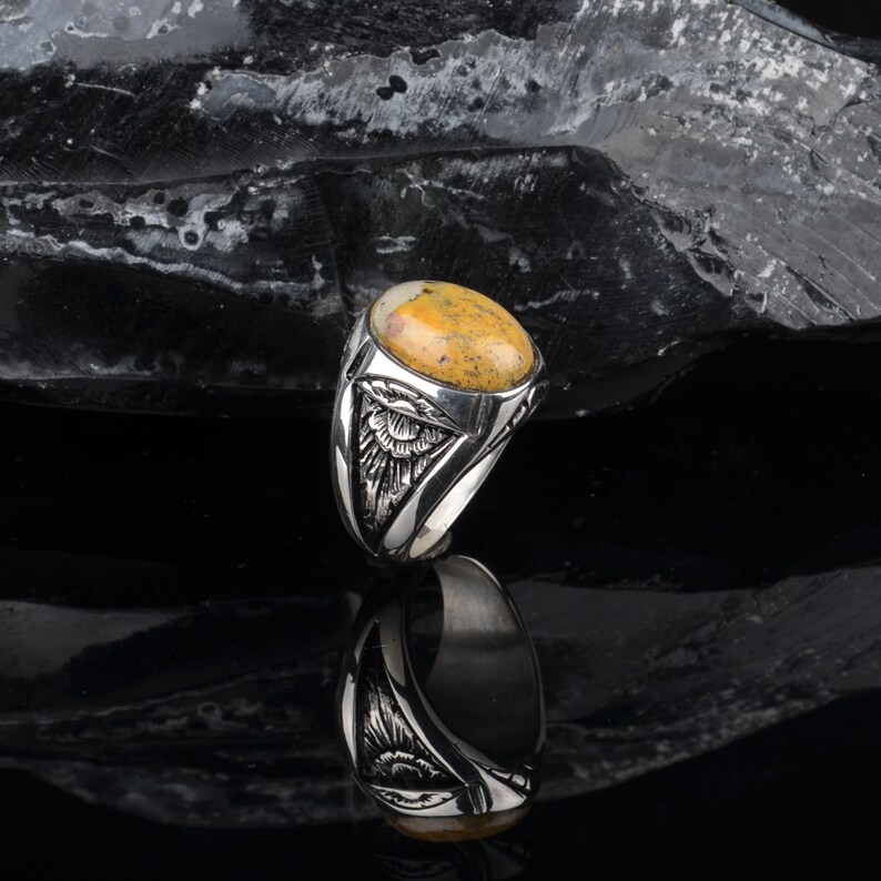 Peut inclure: Une bague en argent avec une pierre ovale jaune et brune sertie dans un entourage en argent d&eacute;taill&eacute;. La bague est sur un fond noir.