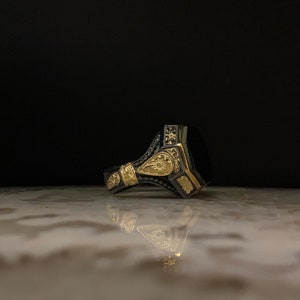 May include: A detailed close-up of a silver ring with gold accents and a large, rectangular black stone. The ring features intricate floral and geometric patterns, with a dark background.