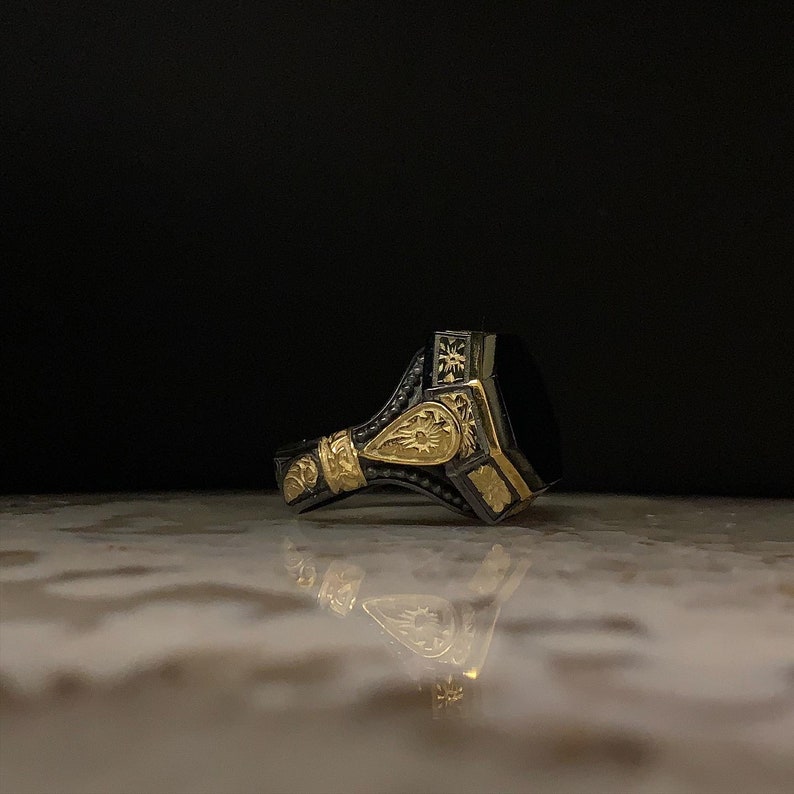 May include: A detailed close-up of a silver ring with gold accents and a large, rectangular black stone. The ring features intricate floral and geometric patterns, with a dark background.