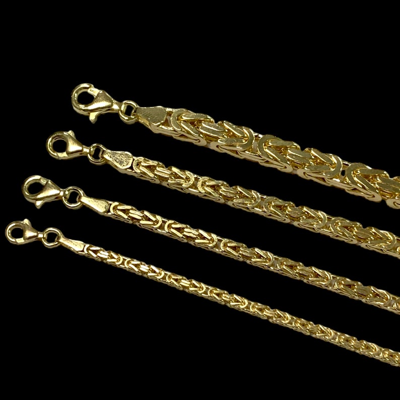 May include: Four gold-colored Byzantine chains of varying thicknesses are displayed against a black background. Each chain features a lobster clasp. The chains are intricately linked, showcasing a classic jewelry design.