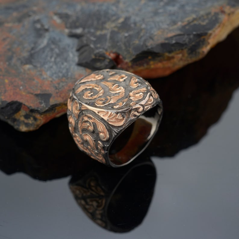 May include: A detailed, two-tone ring with an ornate, swirling pattern. The ring features a dark, possibly oxidized, base metal with raised, gold-colored filigree. The design is symmetrical and covers the entire top surface of the ring.