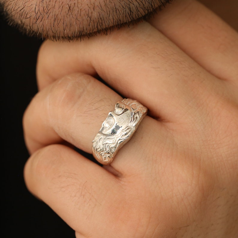 May include: A silver ring with a detailed design of a face. The ring is worn on a person's finger.