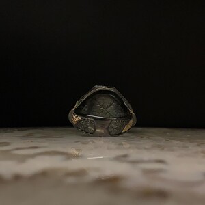 May include: A silver ring with a dark, textured interior and ornate floral detailing. The ring has gold accents on the sides and a geometric pattern inside. The ring is set against a dark background.