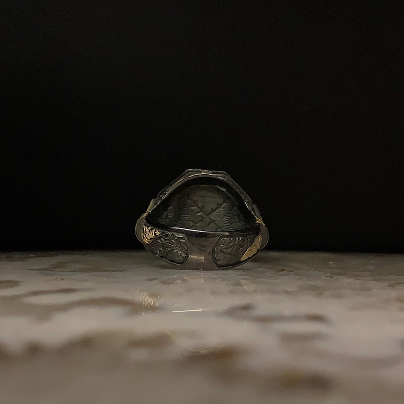 May include: A silver ring with a dark, textured interior and ornate floral detailing. The ring has gold accents on the sides and a geometric pattern inside. The ring is set against a dark background.