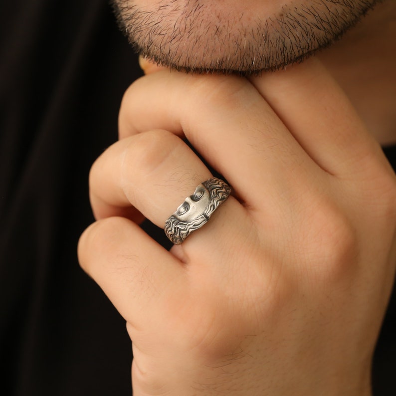 May include: A silver ring with a detailed design of a face with flowing hair. The ring is worn on a person's finger.