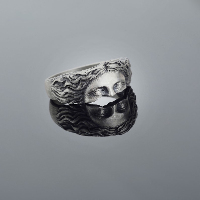 May include: A silver ring with a detailed sculpted design of a woman's face with flowing hair.