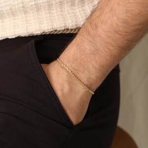 K&ouml;nnte beinhalten: Ein goldfarbenes Kettenarmband. Das Armband wird am Handgelenk getragen, mit einer schwarzen Tasche und einem cremefarbenen Hemd im Hintergrund. Das Armband ist ein zartes Accessoire.