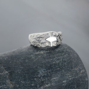 May include: A silver ring with a detailed design of a woman's face. The ring is sitting on a gray rock.