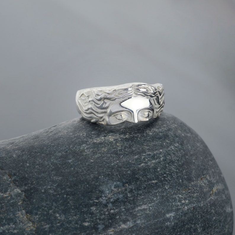May include: A silver ring with a detailed design of a woman's face. The ring is sitting on a gray rock.
