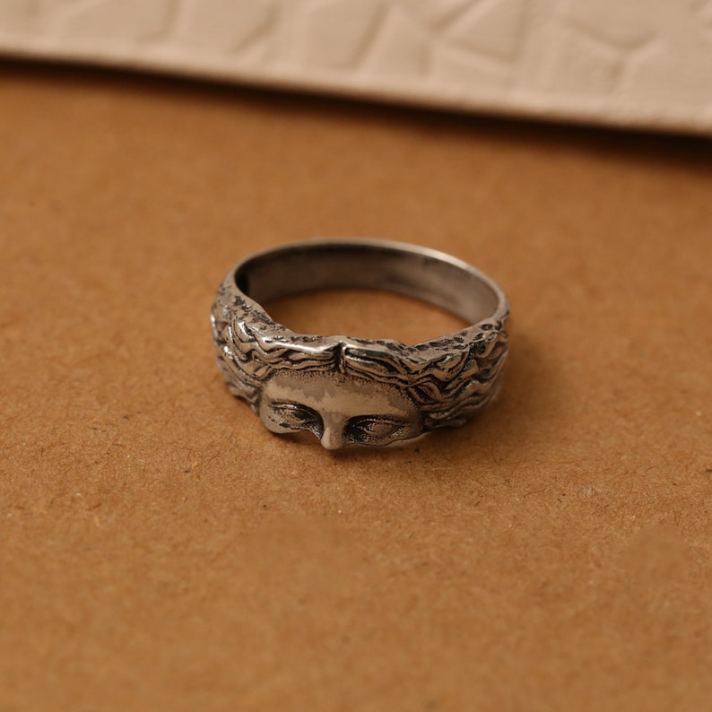 K&ouml;nnte beinhalten: Ein silberfarbener Ring mit detailliertem Gesichtsdesign. Der Ring hat ein strukturiertes Band und ein geformtes Gesicht mit geschlossenen Augen und flie&szlig;endem Haar. Der Ring ist wahrscheinlich aus Metall und ein Schmuckst&uuml;ck.
