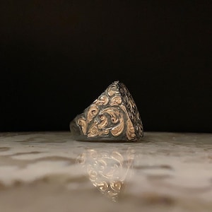 May include: A silver ring with intricate gold filigree detailing. The ring features a dark background and a reflective surface, highlighting the ornate scrollwork and design. The ring's design is a classic signet style.