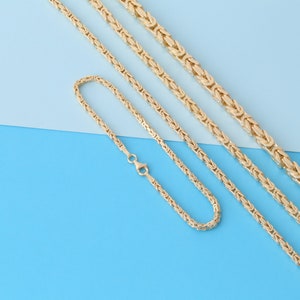 May include: Gold-colored Byzantine chain necklace and bracelet set. The jewelry is displayed on a blue and light blue background. The necklace has a lobster clasp closure. The bracelet is also visible.