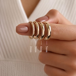 May include: A close-up of five gold-colored hoop rings of varying widths, from 2mm to 5mm, displayed on a finger. The rings have a polished, reflective surface, and the sizes are labeled below each ring.