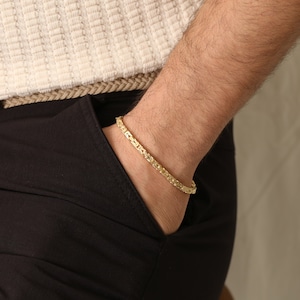 May include: A gold-colored bracelet with a linked chain design. The bracelet is worn on a wrist, with the arm resting in a pocket. The bracelet is approximately 7 inches in length.