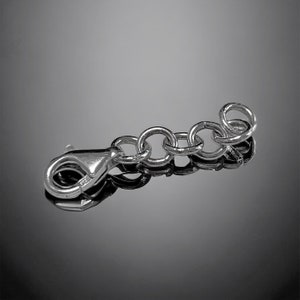 May include: A silver-colored jewelry extender chain with a lobster clasp and several circular links. The chain is designed to add length to necklaces or bracelets. The clasp is closed, and the chain is displayed against a reflective surface.