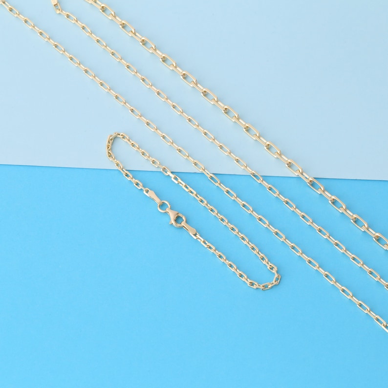 K&ouml;nnte beinhalten: Vier goldfarbene Kettenhalsketten und ein Armband werden vor einem blau und hellblauen Hintergrund gezeigt. Die Ketten haben ein filigranes, verbundenes Design, mit einem Karabinerverschluss am Armband.