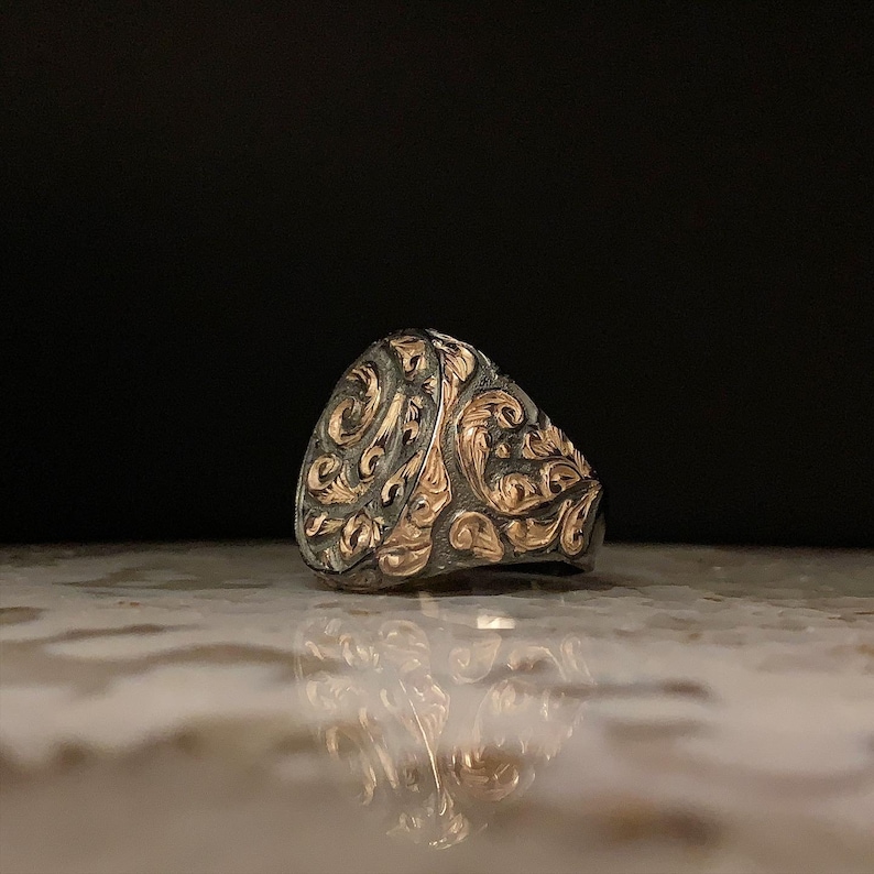May include: A silver ring with intricate gold filigree detailing. The ring features a rounded face with an ornate, swirling pattern. The design is a mix of silver and gold, creating a striking contrast. The ring is likely a piece of jewelry.